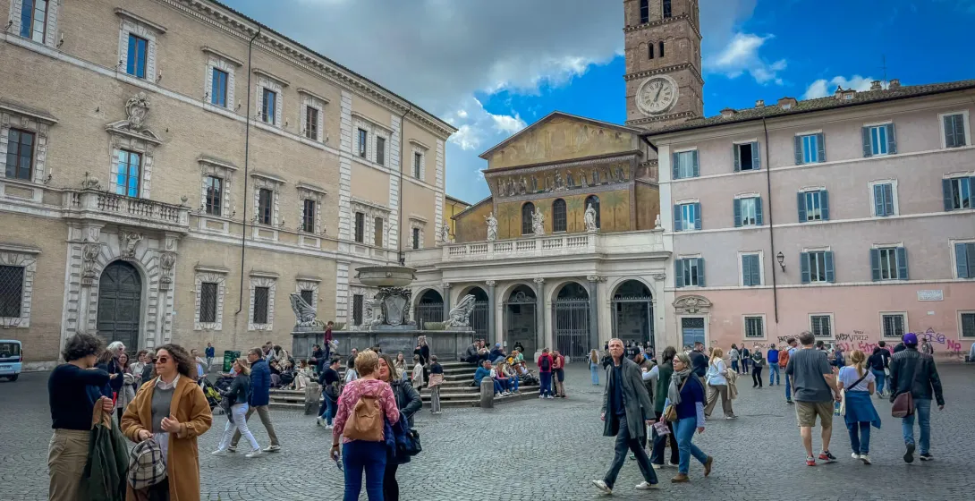 Brukowana Piazza Santa Maria w Trastevere, Rzym, z bazyliką, dzwonnicą, fontanną i spacerującymi ludźmi.