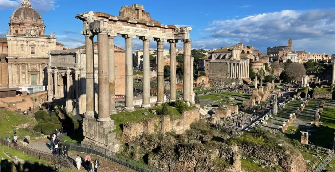 Ruinen des Forum Romanum mit Säulen des Saturn-Tempels, weiteren Tempeln und Besuchern unter blauem Himmel.