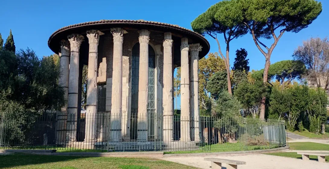 Ancient round Temple of Hercules Victor in Rome featuring Corinthian columns, surrounded by trees and a metal fence.
