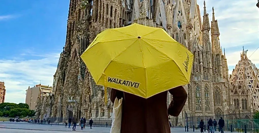 Person holding a yellow Walkative umbrella, facing the intricate spires of Sagrada Família under a blue sky in Barcelona.