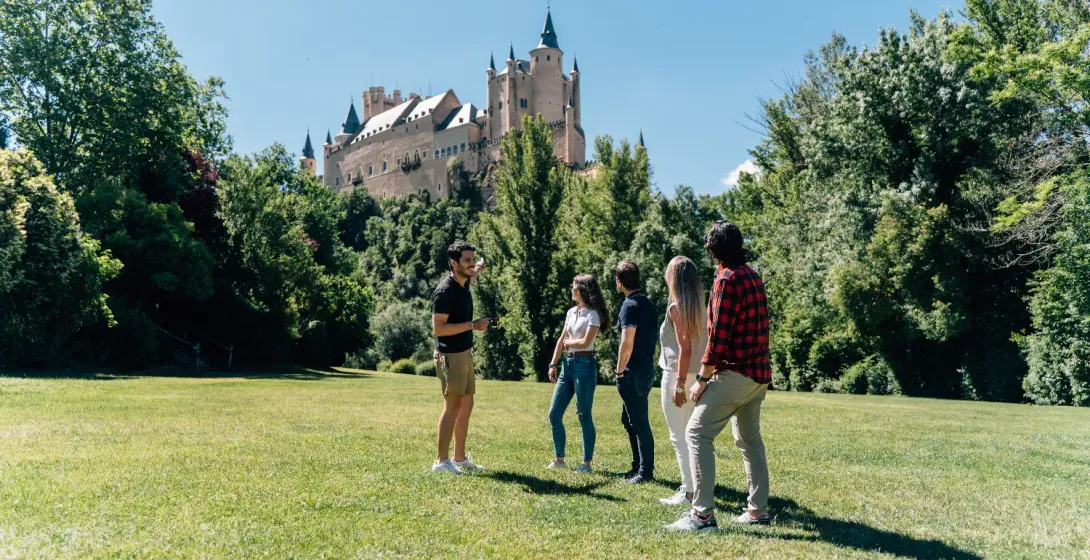 Un guía turístico habla con un grupo pequeño en un parque con el Alcázar de Segovia al fondo.