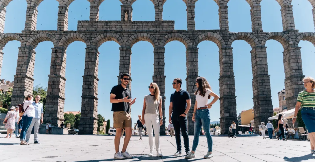 Un guía turístico habla con tres turistas frente al gran y soleado Acueducto de Segovia, España.