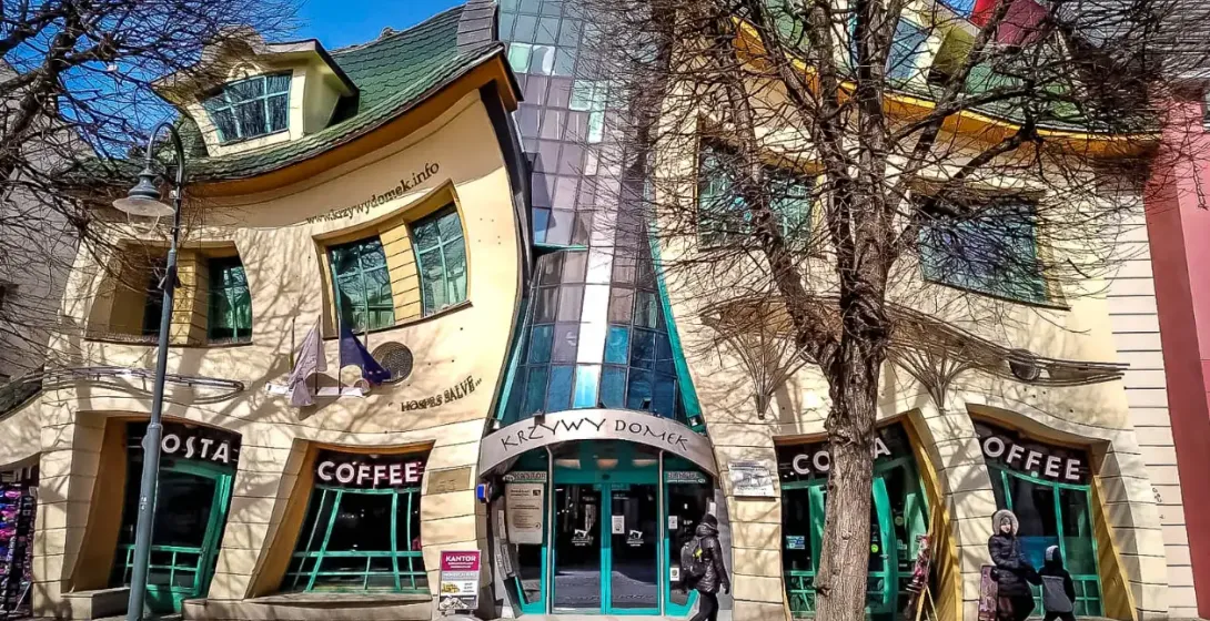 Das Krumme Haus (Krzywy Domek) in Sopot, mit geschwungener Fassade und grünem Dach, Geschäfte im Erdgeschoss, kahle Bäume.