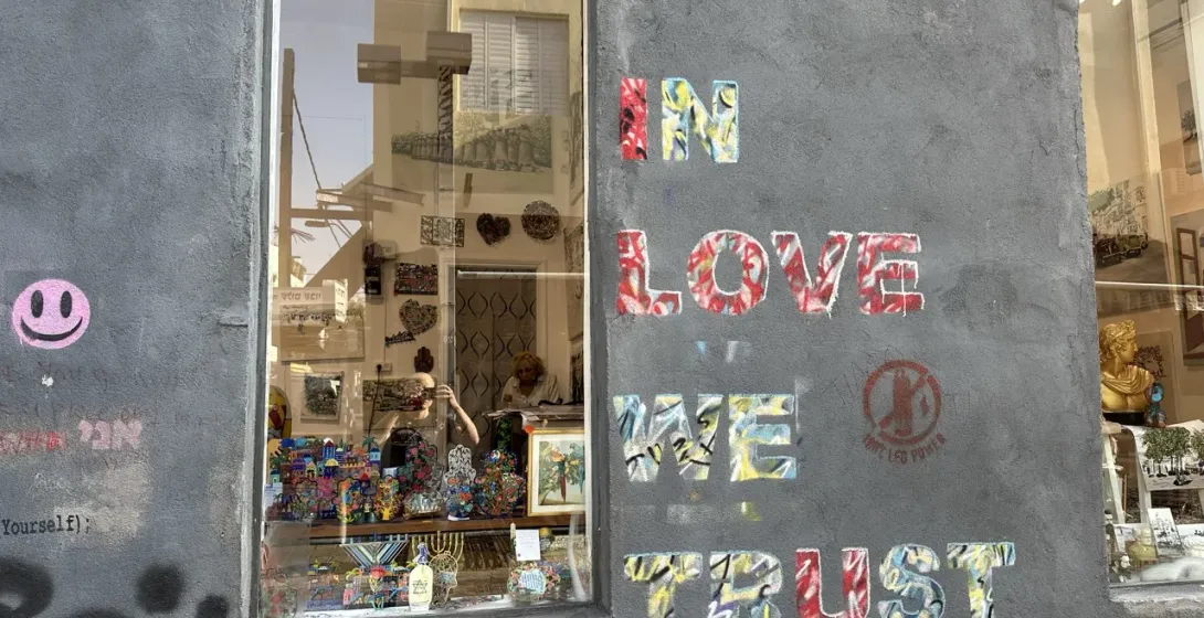 Grey wall with colorful "IN LOVE WE TRUST" graffiti, a pink smiley face, and a shop window reflecting buildings.