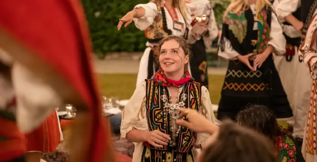 Młoda kobieta w haftowanym tradycyjnym stroju albańskim i czerwonej chuście uśmiecha się, z innymi osobami w tle.