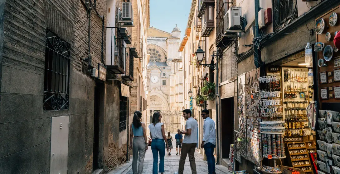 Cuatro jóvenes caminan por un callejón estrecho en Toledo, con la catedral al fondo y una tienda de recuerdos a la derecha.