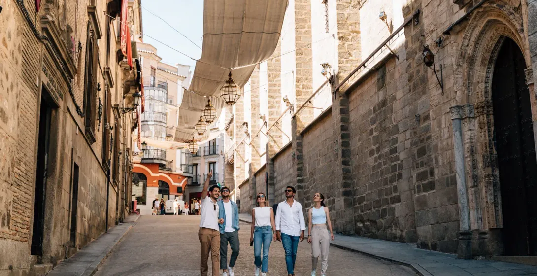 Un grupo de cinco jóvenes pasea por una estrecha calle de piedra en Toledo, a la sombra de los toldos tradicionales.