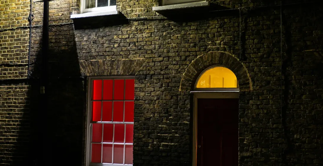 Edificio de ladrillo en una calle oscura de noche, iluminado por una farola. Una ventana brilla con una luz roja.