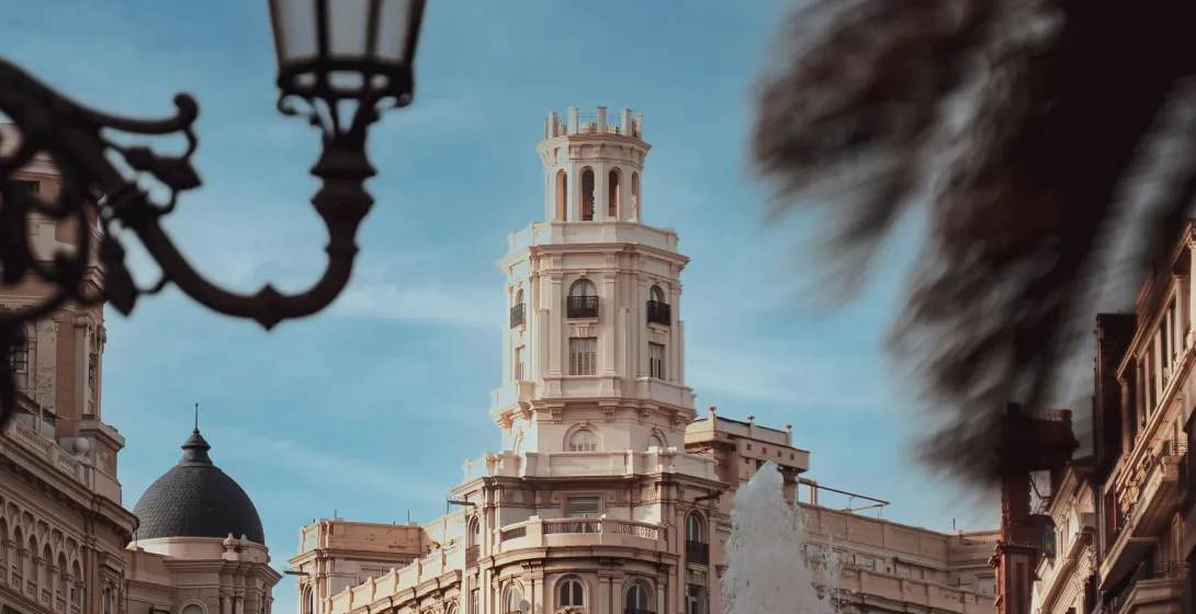Ornate streetlamp frames a distant historic building with a tower, a fountain's spray, and palm fronds.