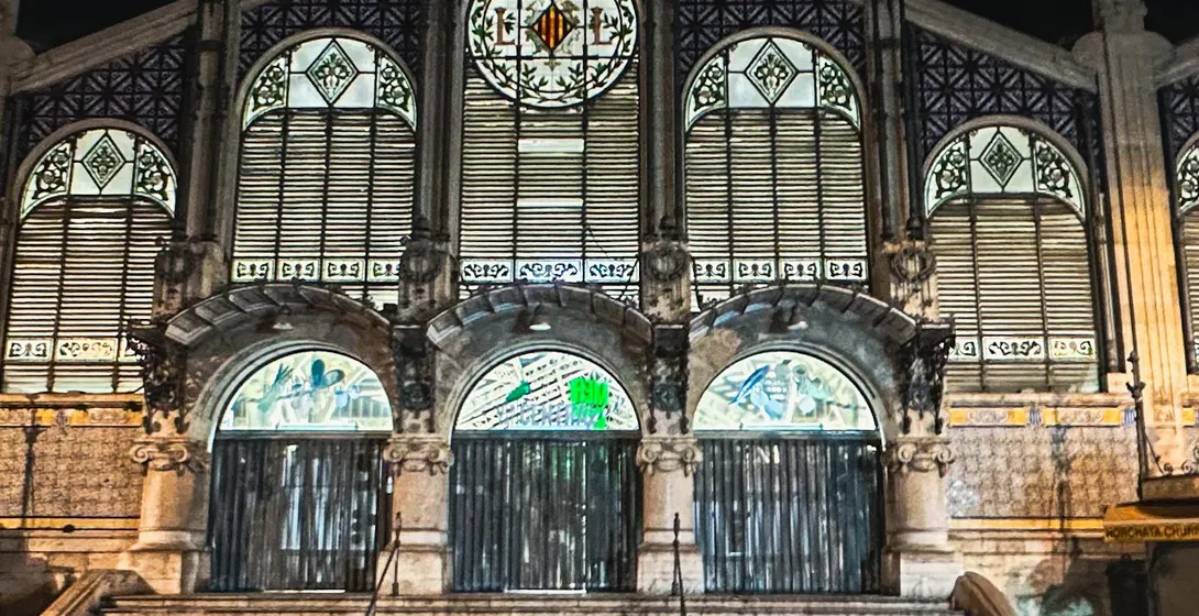 Illuminated Art Nouveau facade of Valencia Central Market at night, featuring arched entrances, steps, and intricate stain...