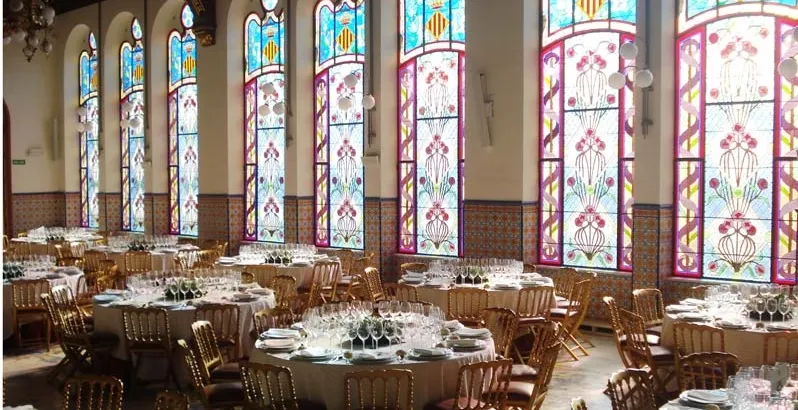 A grand hall with tables set for a banquet, illuminated by tall, colorful stained-glass windows.