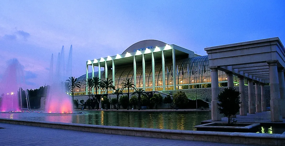 The Palau de la Música in Valencia at dusk, its glass facade lit up, with colourful fountains in the foreground.