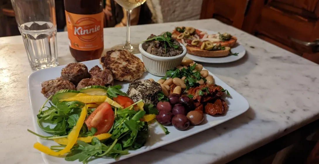 Maltese platter with sausage, bigilla dip, olives, and cheeselet, served with a bottle of Kinnie soft drink.