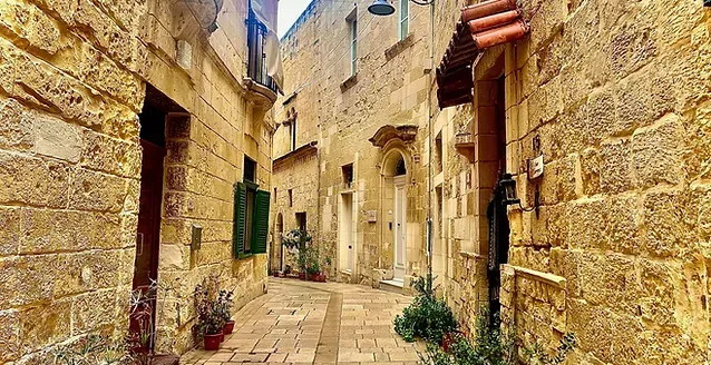A narrow stone street in an old town, flanked by tall buildings with balconies, green shutters, and ornate street lamps.