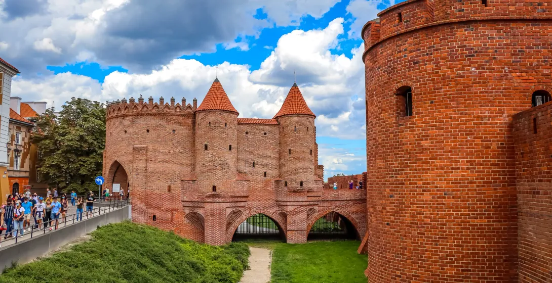 Barbacana de ladrillo rojo con dos torres cónicas y pasajes arqueados. Césped verde y un camino conducen a ella, con gente...