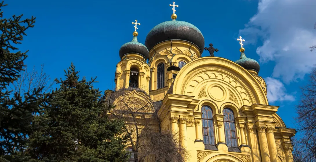 Gelbe orthodoxe Kirche mit dunklen Kuppeln und goldenen Kreuzen vor blauem Himmel, umgeben von grünen Bäumen.
