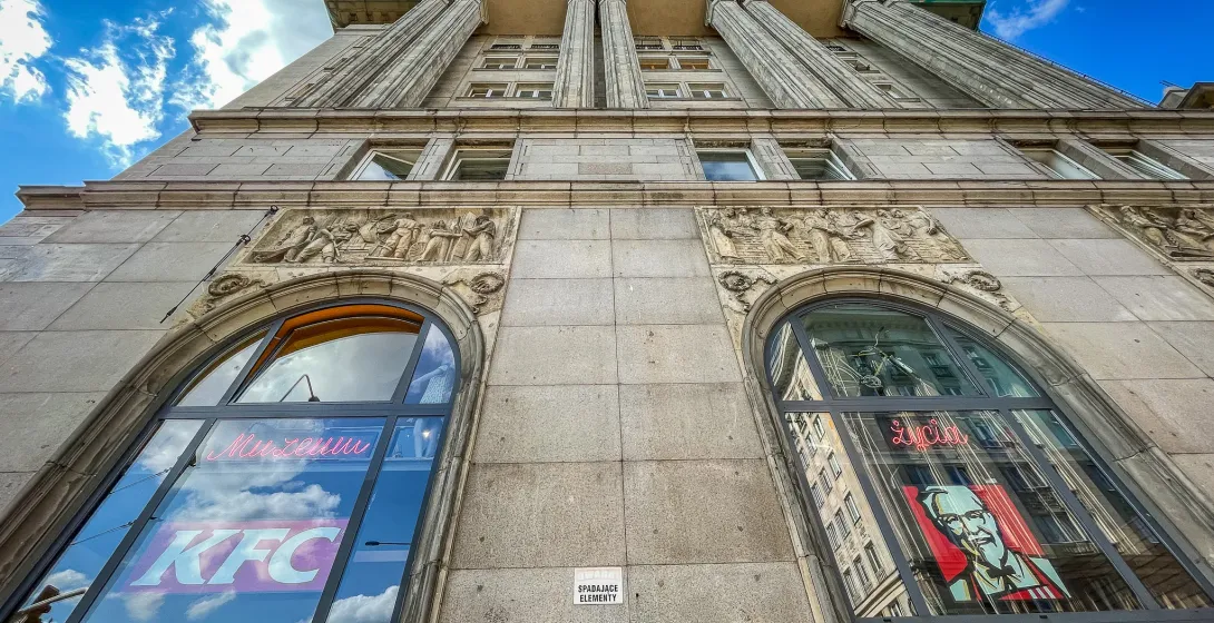 Untersicht eines grauen Steingebäudes mit klassischen Säulen und Reliefschnitzereien. Große Bogenfenster zeigen Reflexione...