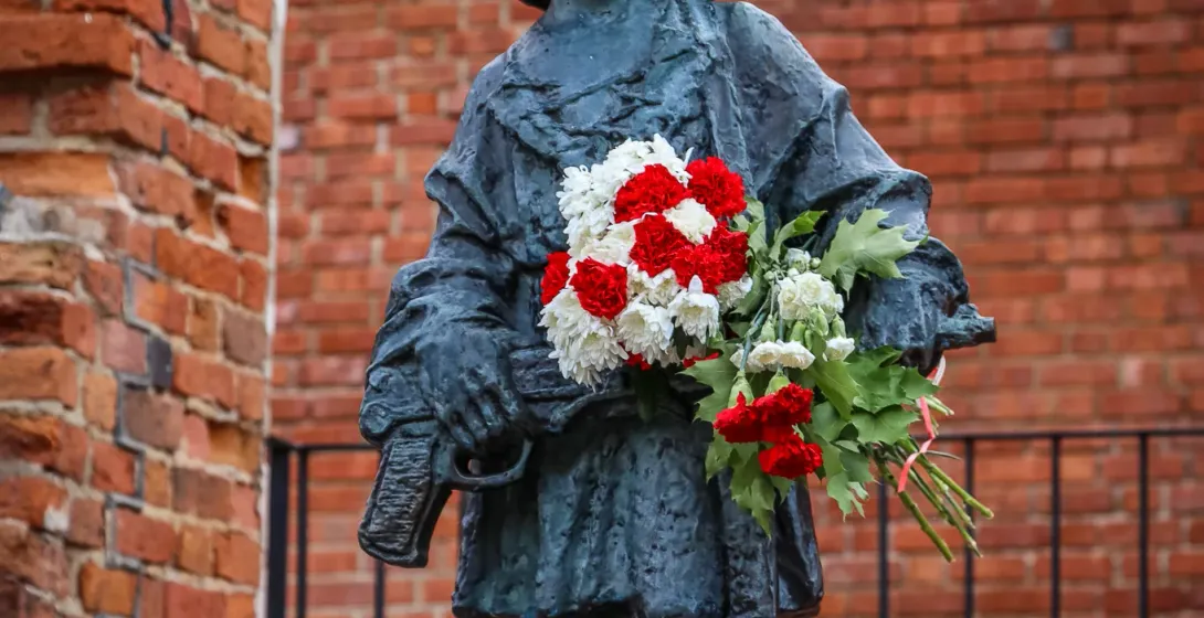 Bronzestatue eines Kinder-Soldaten mit Helm und rot-weißen Blumen vor einer Backsteinmauer.