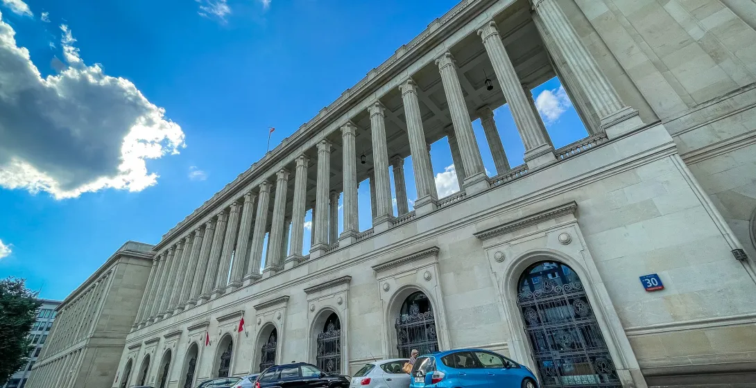 Large, imposing building with a long colonnade and parked cars under a bright blue sky in Warsaw.