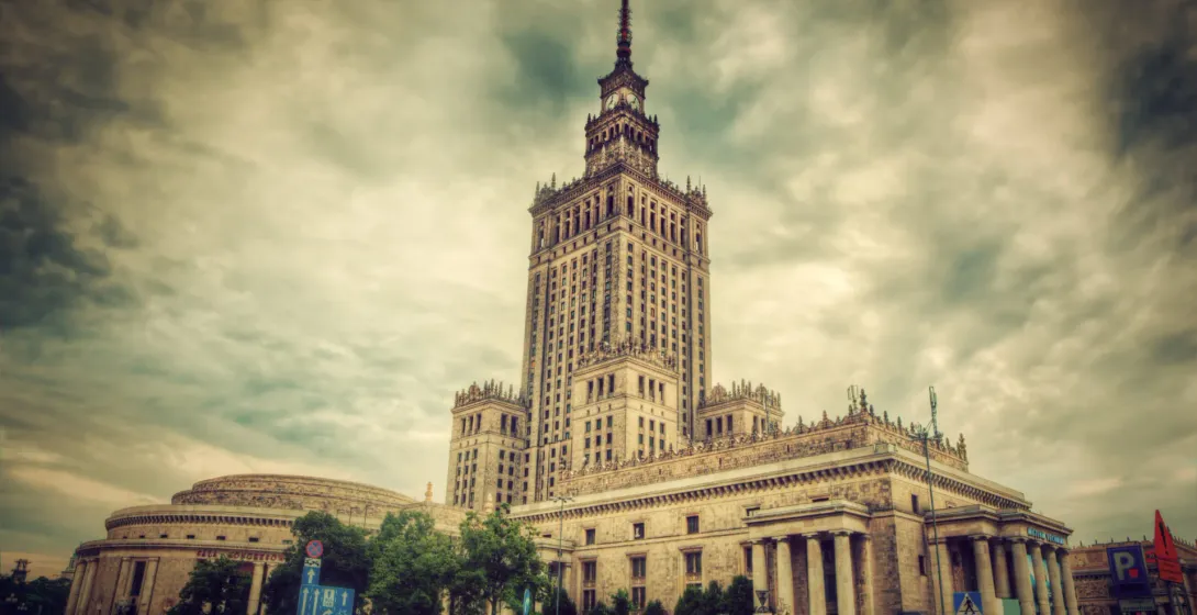 The Palace of Culture and Science in Warsaw, a tall, ornate building with a spire, stands against a cloudy, dramatic sky.