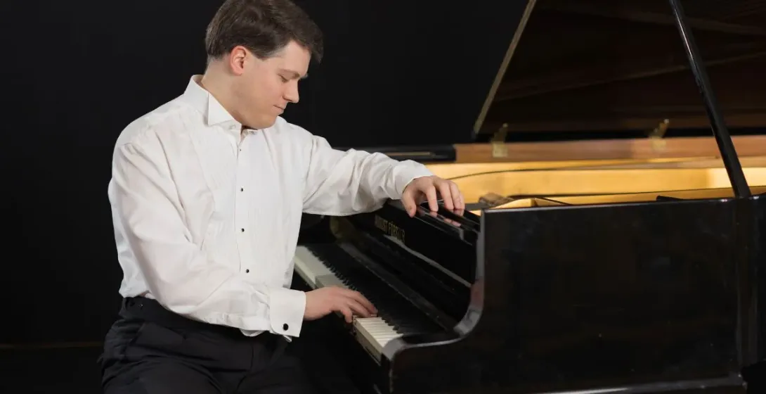 A male pianist in a white tuxedo shirt and black trousers plays a black grand piano against a dark background.