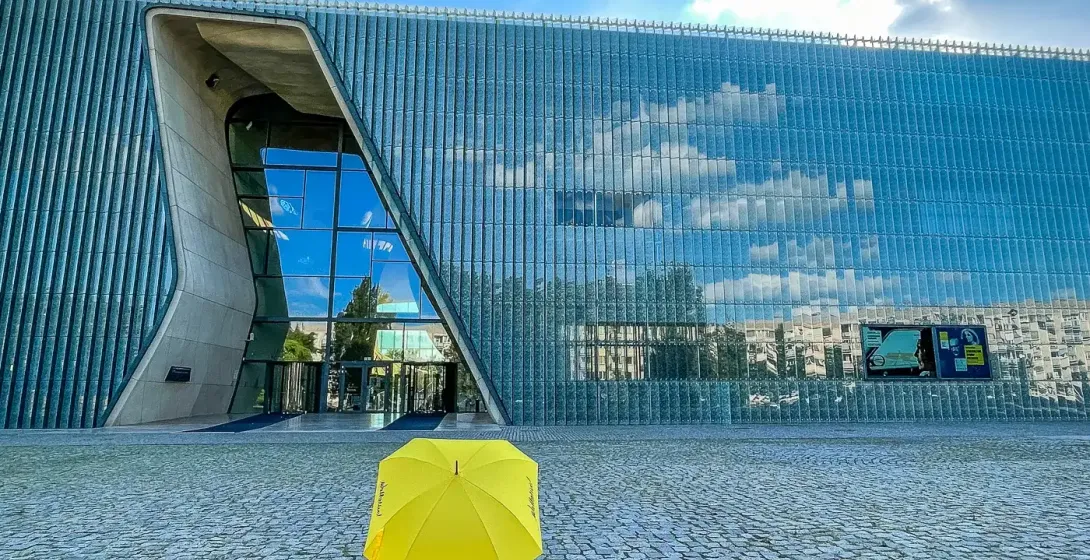 Paraguas amarillo Walkative en plaza adoquinada frente al moderno edificio de cristal del Museo POLIN en Varsovia.