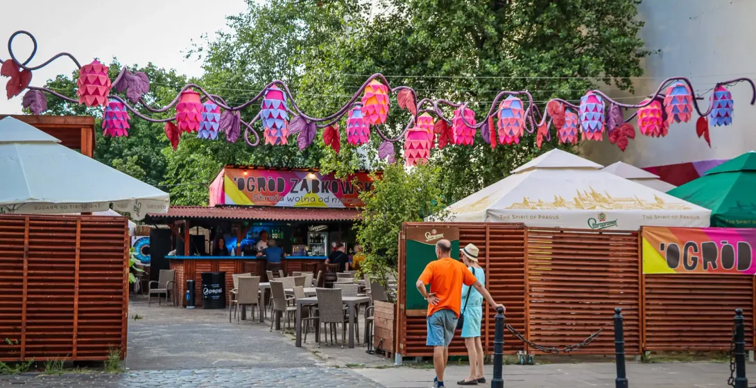 Outdoor bar area under colorful hop-shaped decorations, with tables, chairs, and two people standing on a cobblestone street.