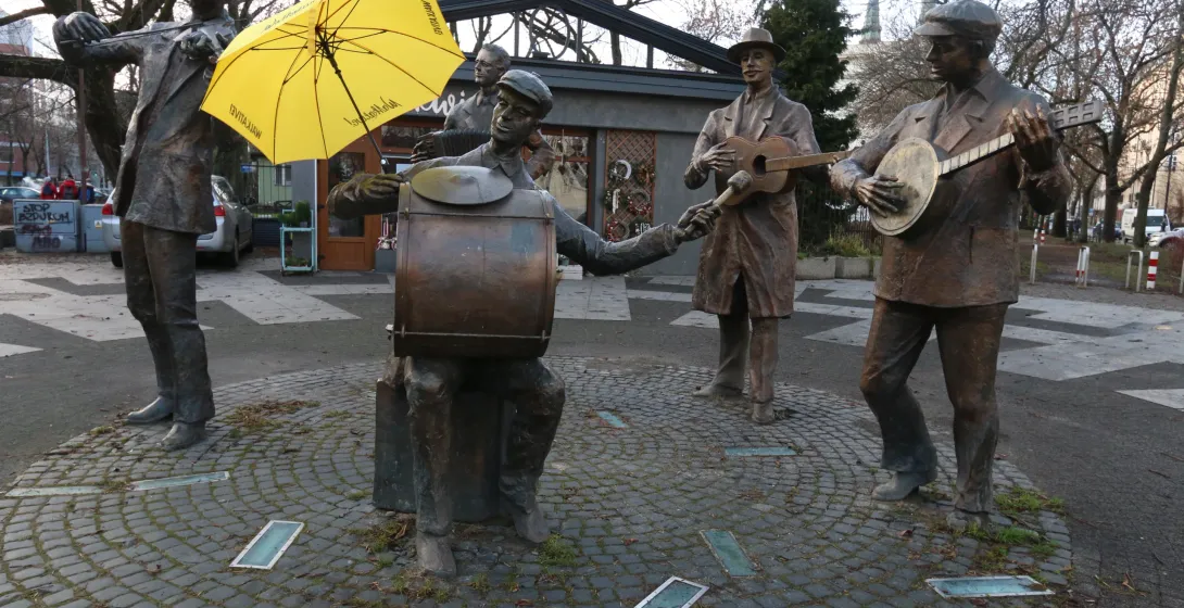Bronze statue of five street musicians in Warsaw's Praga district, one holding a yellow umbrella, on a cobblestone circle.
