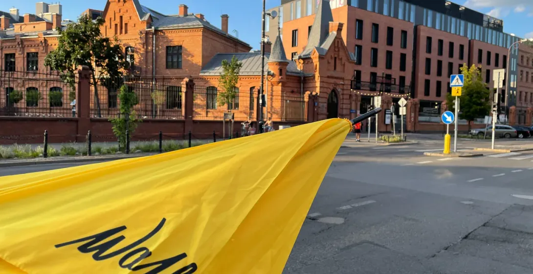 A yellow Walkative umbrella in the foreground, with historical brick buildings, a modern building, and a street in Warsaw'...