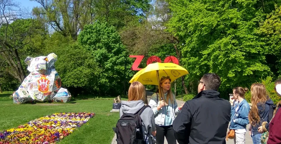 Tour guide with yellow umbrella speaking to a group on a paved path, next to a flower bed and green trees. A ZOO sign and ...