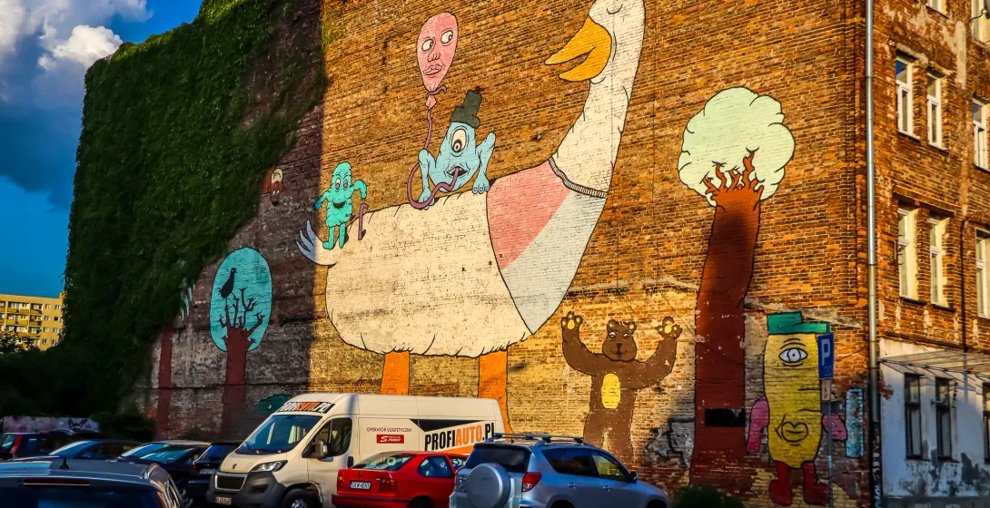 Colorful mural on a brick building in Warsaw, showing a large goose, whimsical figures, and a bear, with parked cars below.