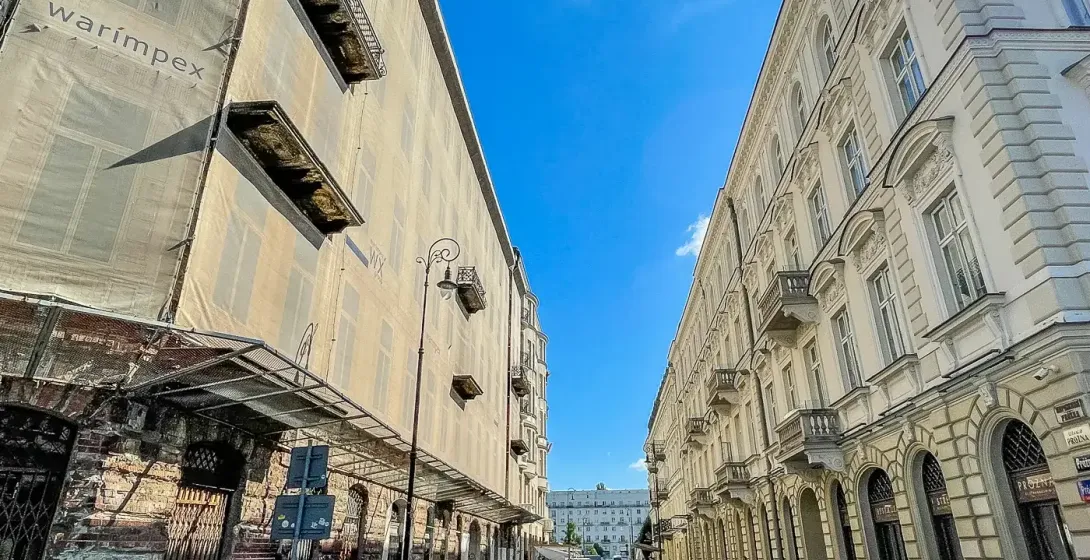 Calle adoquinada entre un edificio andamiado con 'warimpex' y un edificio histórico ornamentado con balcones bajo cielo azul.