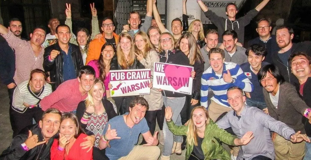 Eine große Gruppe junger Erwachsener lächelt und posiert mit „Pub Crawl Warsaw“- und „Wir lieben Warschau“-Schildern in st...