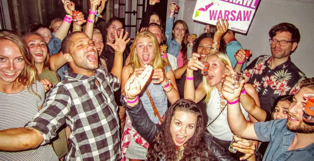 Gruppe junger Erwachsener lachend und lächelnd, mit Schnapsgläsern, einer hält ein 'PUB CRAWL WARSAW' Schild.