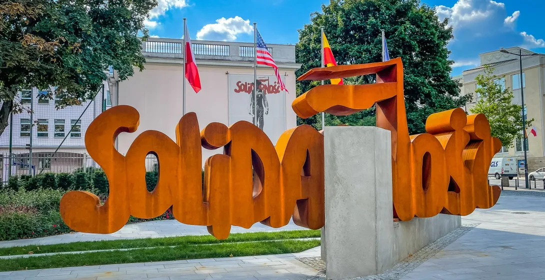 Rostfarbene Skulptur mit 'Solidarność' in Warschau. Polnische und US-Flaggen, Bäume und ein weißes Gebäude im Hintergrund.