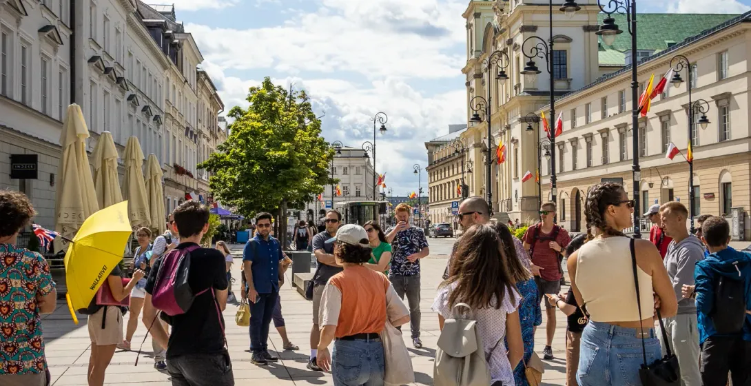 Un grupo de personas reunidas en una calle adoquinada de Varsovia con una iglesia histórica y edificios de fondo bajo un c...