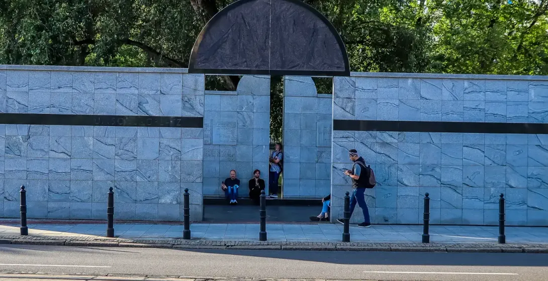 Muro conmemorativo de mármol gris con arco semicircular oscuro, personas de pie y sentadas, vías de tranvía y calle en pri...