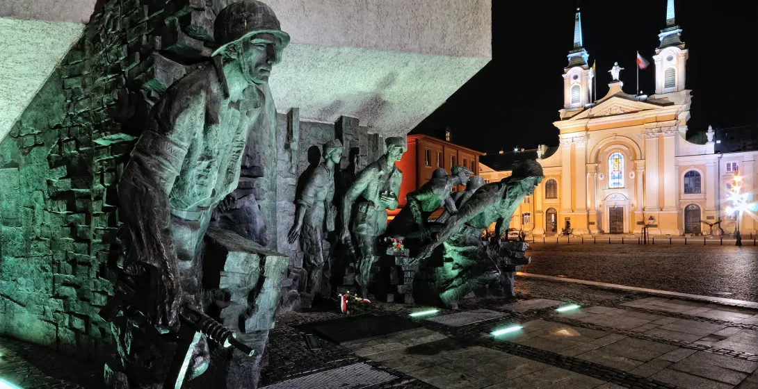 Bronzemonument mit Soldaten des Warschauer Aufstands, nachts grün beleuchtet, im Hintergrund eine hell erleuchtete Kirche.