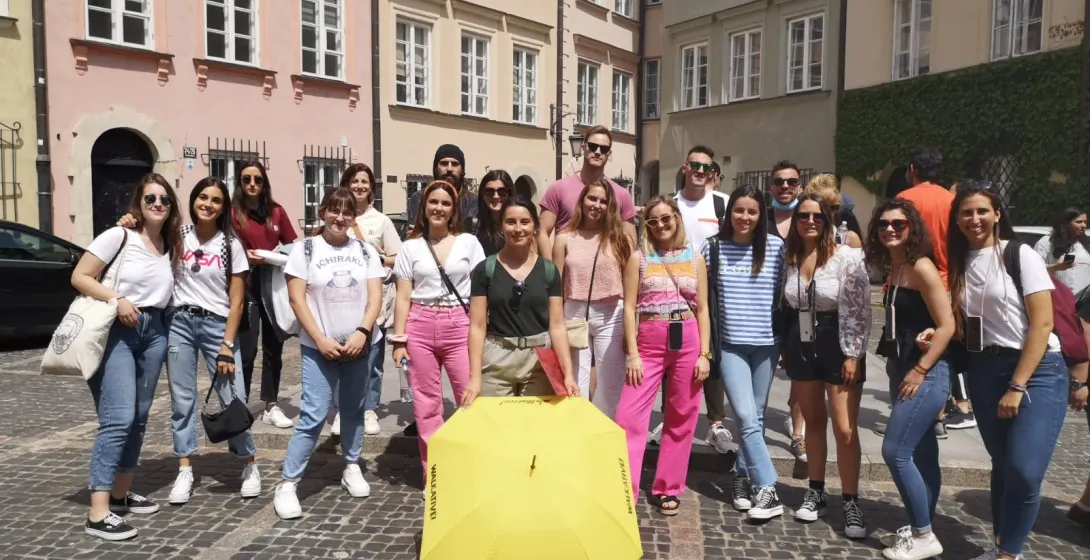 Un grupo de jóvenes posa en una calle empedrada con un paraguas amarillo de Walkative. Edificios históricos coloridos al f...
