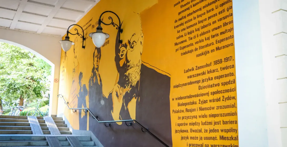 Mural amarillo de Ludwik Zamenhof con texto, junto a escaleras de cemento con pasamanos y farolas.