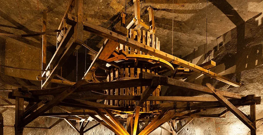 A large, historical wooden horse-powered hoisting machine inside the Wieliczka Salt Mine, with salt rock walls behind it.