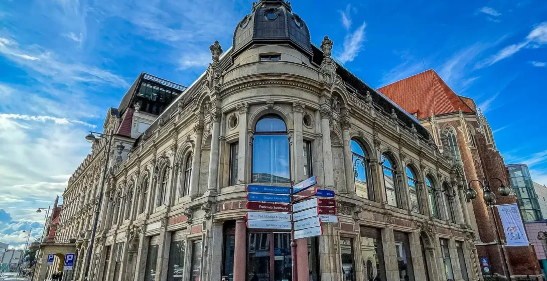 Ornate historic building with a domed corner tower and large windows on a cobblestone street in Wrocław. A street signpost...