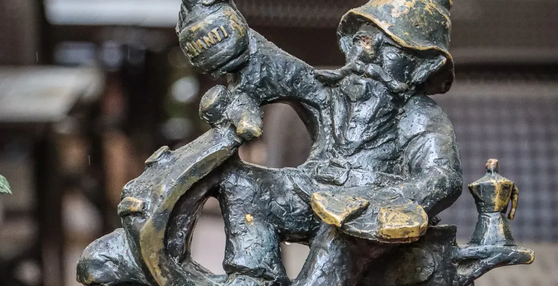 Bronze statue of a Wroclaw dwarf riding a scooter. The dwarf wears a pointy hat and holds a bottle of Chianti. A moka pot ...