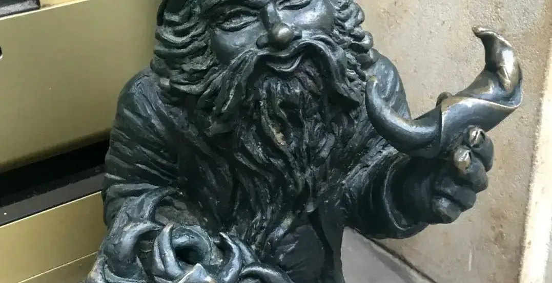 Bronze dwarf statue with a pointed hat and long beard, holding a tray with knots and a horn-shaped object, next to a build...