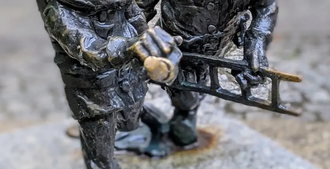 Dwie ciemne, brązowe figurki krasnali z Wrocławia w złotych hełmach, jeden z brodą trzymający kilof, drugi drabinę.