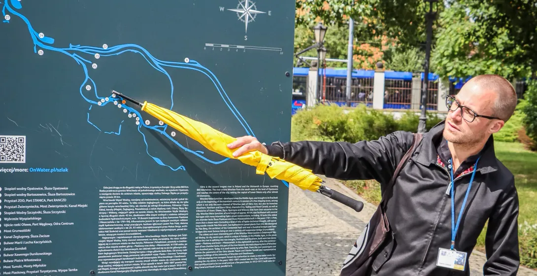 Mann zeigt mit gelbem Regenschirm auf eine Informationstafel mit Karte des Breslauer Wasserstraßensystems an einem sonnige...