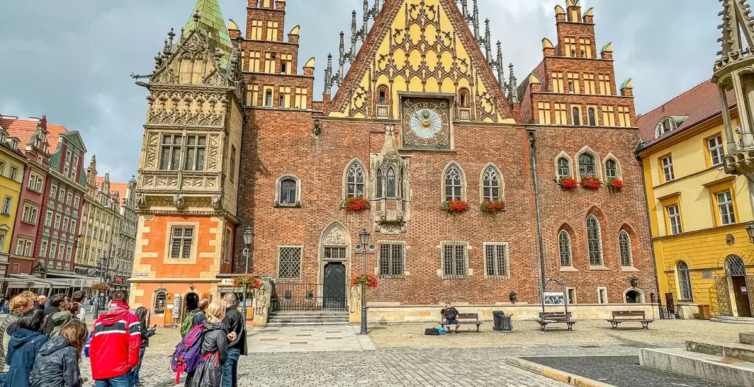 Wrocławski Ratusz, duży gotycki budynek z cegły z wysoką wieżą zegarową, stoi na brukowanym Rynku z grupą ludzi.