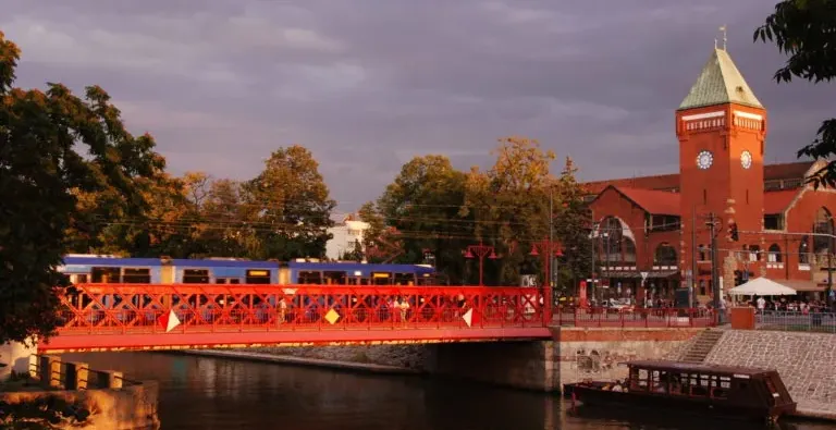 Rote Metallbrücke über einem Fluss, ein blauer Zug fährt darüber, daneben eine rote Backstein-Markthalle mit Uhrturm bei S...