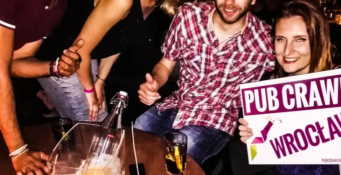Four smiling young adults at a table with drinks and shots, one holding a 'PUB CRAWL WROCŁAW' sign.
