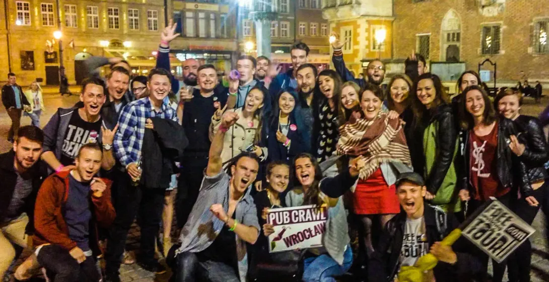 A large group of young adults smiling and posing for a photo on a street at night. One person holds a 'Pub Crawl Wrocław' ...